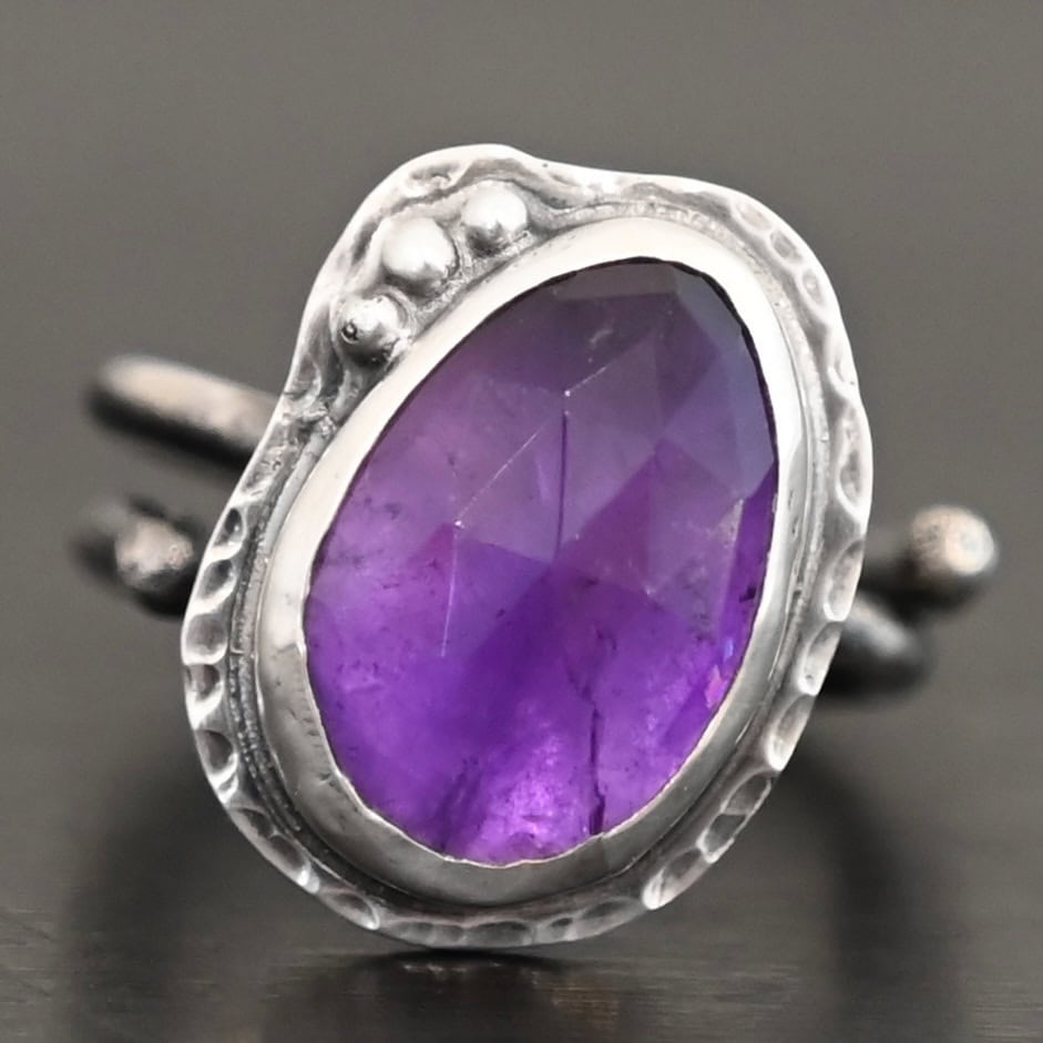 Photo of the "Let It Blossom" Ring. Ring is made out of sterling silver and amethyst. The amethyst is an oval shape. It is a dark purple color. It is set in sterling silver. There is hammered sterling silver around the stone setting as well in an oval shape. On the hammered sterling silver are three silver balls on the left side of the amethyst. They are right next to one another and follow the curve of the amethyst. Ring has one band that does a loop around so that it looks like two bands. Ring is sitting against a dark background.