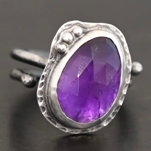 Photo of the "Let It Blossom" Ring. Ring is made out of sterling silver and amethyst. The amethyst is an oval shape. It is a dark purple color. It is set in sterling silver. There is hammered sterling silver around the stone setting as well in an oval shape. On the hammered sterling silver are three silver balls on the left side of the amethyst. They are right next to one another and follow the curve of the amethyst. Ring has one band that does a loop around so that it looks like two bands. Ring is sitting against a dark background.
