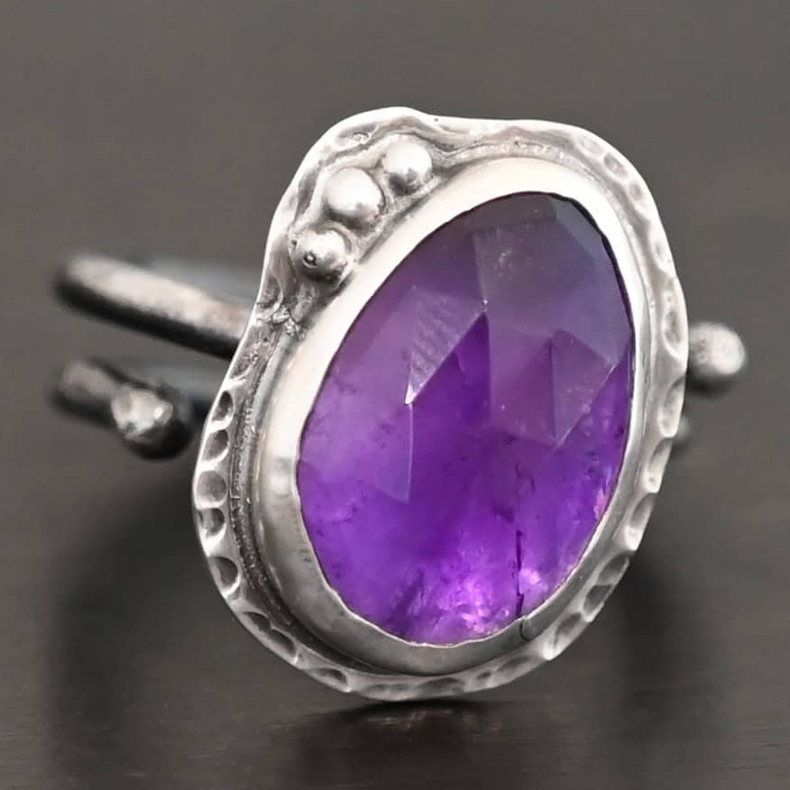 Photo of the "Let It Blossom" Ring. Ring is made out of sterling silver and amethyst. The amethyst is an oval shape. It is a dark purple color. It is set in sterling silver. There is hammered sterling silver around the stone setting as well in an oval shape. On the hammered sterling silver are three silver balls on the left side of the amethyst. They are right next to one another and follow the curve of the amethyst. Ring has one band that does a loop around so that it looks like two bands. Ring is sitting against a dark background.