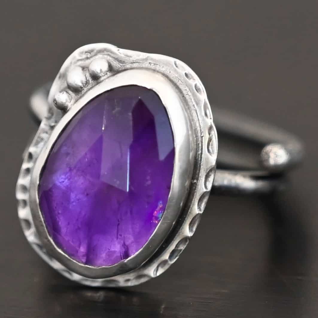 Photo of the "Let It Blossom" Ring. Ring is made out of sterling silver and amethyst. The amethyst is an oval shape. It is a dark purple color. It is set in sterling silver. There is hammered sterling silver around the stone setting as well in an oval shape. On the hammered sterling silver are three silver balls on the left side of the amethyst. They are right next to one another and follow the curve of the amethyst. Ring has one band that does a loop around so that it looks like two bands. Ring is sitting against a dark background.