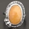 Close up of the "Marigold" Ring. Made out of sterling silver and sapphire. The sapphire is an oval shape and rosecut, and it is a dark yellow color. In the bottom left corner on the hammered sterling silver surrounding the ring are three silver balls. They are slightly curved and set next to one another so that they follow the curve of the sapphire. Ring is sitting against a dark background.