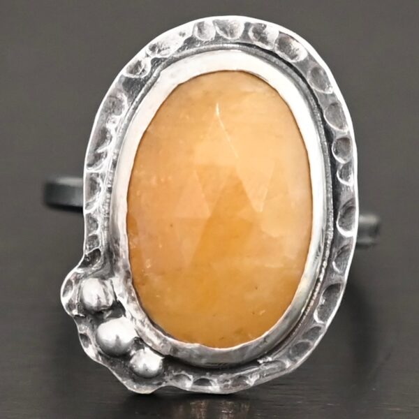 Close up of the "Marigold" Ring. Made out of sterling silver and sapphire. The sapphire is an oval shape and rosecut, and it is a dark yellow color. In the bottom left corner on the hammered sterling silver surrounding the ring are three silver balls. They are slightly curved and set next to one another so that they follow the curve of the sapphire. Ring is sitting against a dark background.