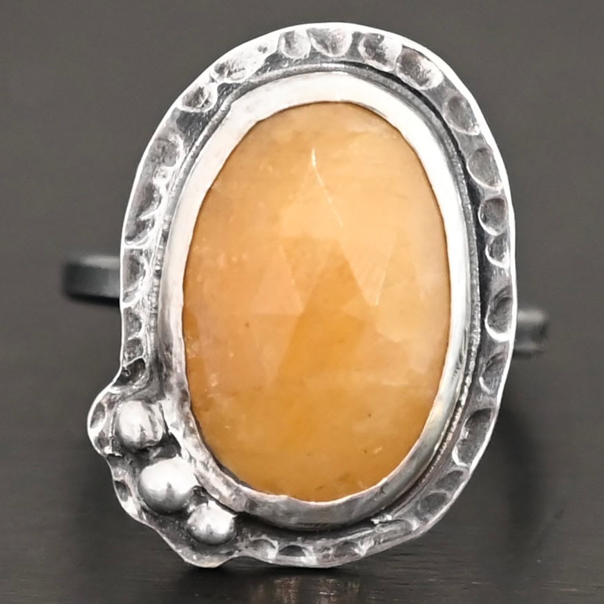 Marigold Ring Close up of the "Marigold" Ring. Made out of sterling silver and sapphire. The sapphire is an oval shape and rosecut, and it is a dark yellow color. In the bottom left corner on the hammered sterling silver surrounding the ring are three silver balls. They are slightly curved and set next to one another so that they follow the curve of the sapphire. Ring is sitting against a dark background.