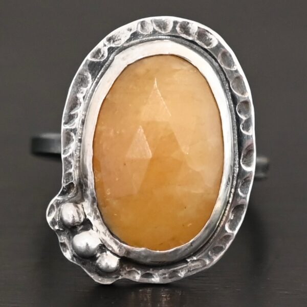 Close up of the "Marigold" Ring. Made out of sterling silver and sapphire. The sapphire is an oval shape and rosecut, and it is a dark yellow color. In the bottom left corner on the hammered sterling silver surrounding the ring are three silver balls. They are slightly curved and set next to one another so that they follow the curve of the sapphire. Ring is sitting against a dark background.