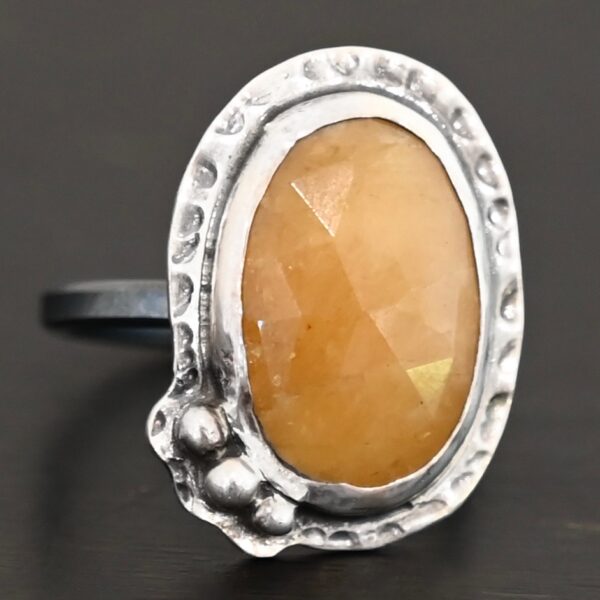 Close up of the "Marigold" Ring. Made out of sterling silver and sapphire. The sapphire is an oval shape and rosecut, and it is a dark yellow color. In the bottom left corner on the hammered sterling silver surrounding the ring are three silver balls. They are slightly curved and set next to one another so that they follow the curve of the sapphire. Ring is sitting against a dark background.