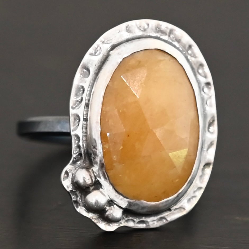 Marigold Ring Close up of the "Marigold" Ring. Made out of sterling silver and sapphire. The sapphire is an oval shape and rosecut, and it is a dark yellow color. In the bottom left corner on the hammered sterling silver surrounding the ring are three silver balls. They are slightly curved and set next to one another so that they follow the curve of the sapphire. Ring is sitting against a dark background.