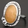 Close up of the "Marigold" Ring. Made out of sterling silver and sapphire. The sapphire is an oval shape and rosecut, and it is a dark yellow color. In the bottom left corner on the hammered sterling silver surrounding the ring are three silver balls. They are slightly curved and set next to one another so that they follow the curve of the sapphire. Ring is sitting against a dark background.
