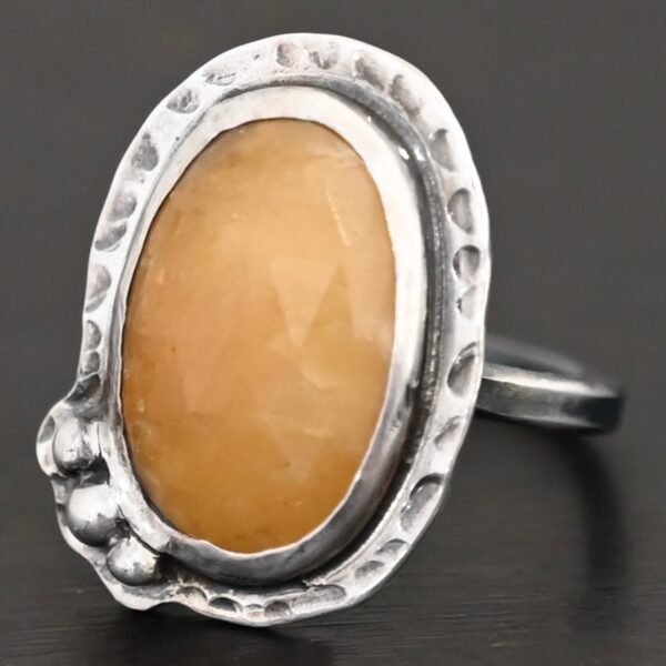Close up of the "Marigold" Ring. Made out of sterling silver and sapphire. The sapphire is an oval shape and rosecut, and it is a dark yellow color. In the bottom left corner on the hammered sterling silver surrounding the ring are three silver balls. They are slightly curved and set next to one another so that they follow the curve of the sapphire. Ring is sitting against a dark background.