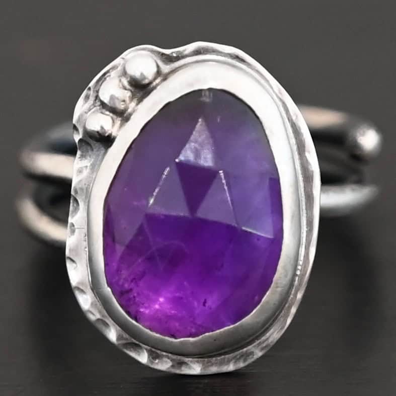 Photo of the "A Life Worthwhile" Ring. Ring is made out of sterling silver and amethyst. The amethyst is an oval shape. It is a dark purple color. It is set in sterling silver. There is hammered sterling silver around the stone setting as well in an oval shape. On the hammered sterling silver are three silver balls on the bottom right side of the amethyst. They are right next to one another and follow the curve of the amethyst. Ring has one band that does a loop around so that it looks like two bands. Ring is sitting against a dark background.