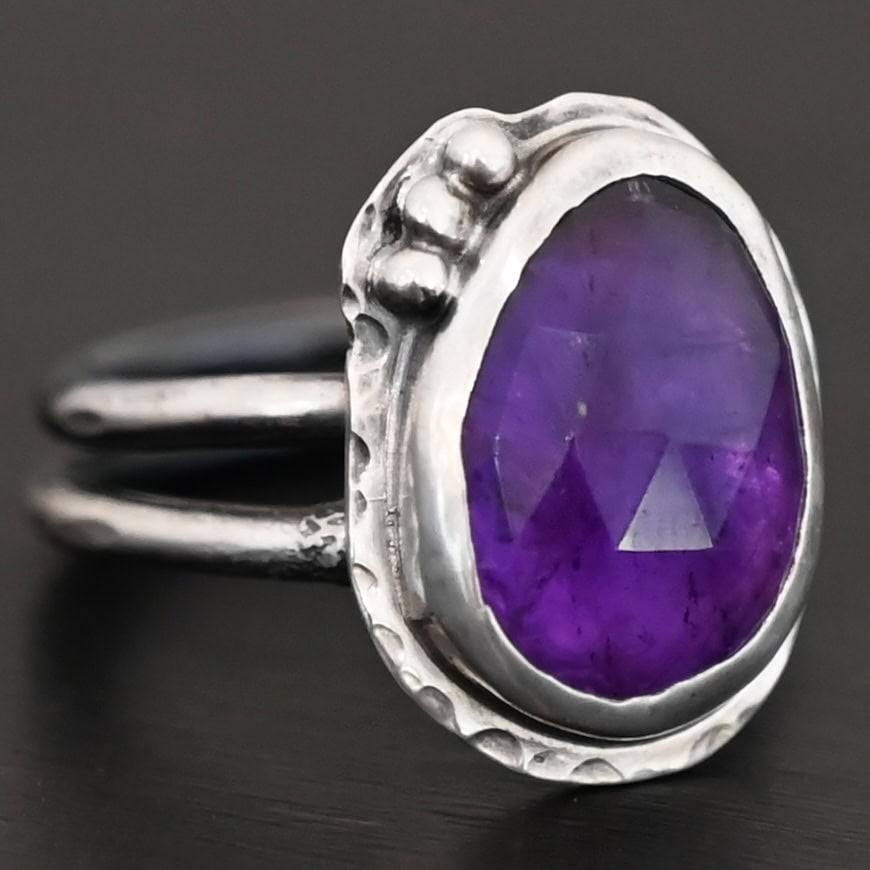 Photo of the "A Life Worthwhile" Ring. Ring is made out of sterling silver and amethyst. The amethyst is an oval shape. It is a dark purple color. It is set in sterling silver. There is hammered sterling silver around the stone setting as well in an oval shape. On the hammered sterling silver are three silver balls on the bottom right side of the amethyst. They are right next to one another and follow the curve of the amethyst. Ring has one band that does a loop around so that it looks like two bands. Ring is sitting against a dark background.