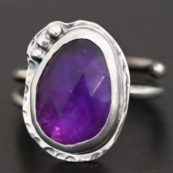 Photo of the "A Life Worthwhile" Ring. Ring is made out of sterling silver and amethyst. The amethyst is an oval shape. It is a dark purple color. It is set in sterling silver. There is hammered sterling silver around the stone setting as well in an oval shape. On the hammered sterling silver are three silver balls on the bottom right side of the amethyst. They are right next to one another and follow the curve of the amethyst. Ring has one band that does a loop around so that it looks like two bands. Ring is sitting against a dark background.