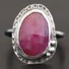 Photo of "Everything Is Rosy" Ring. Made out of sterling silver and sapphire. The sapphire is an oval shape and a dark pink, almost magenta color. It is a rose cut sapphire that is set in silver. Going around the silver setting is more hammered sterling silver in an oval shape. There are three silver balls on the top left of the oval stone setting on the hammered silver. They are right next to one another, and curve so that they go along with the curve of the sapphire. Ring has one band. Ring is sitting against a dark background.