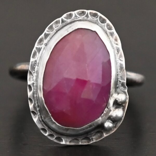 Photo of "Everything Is Rosy" Ring. Made out of sterling silver and sapphire. The sapphire is an oval shape and a dark pink, almost magenta color. It is a rose cut sapphire that is set in silver. Going around the silver setting is more hammered sterling silver in an oval shape. There are three silver balls on the top left of the oval stone setting on the hammered silver. They are right next to one another, and curve so that they go along with the curve of the sapphire. Ring has one band. Ring is sitting against a dark background.
