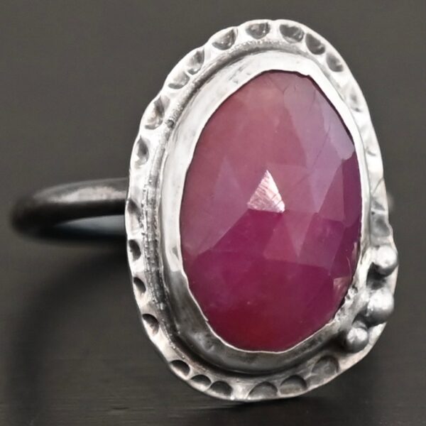 Photo of "Everything Is Rosy" Ring. Made out of sterling silver and sapphire. The sapphire is an oval shape and a dark pink, almost magenta color. It is a rose cut sapphire that is set in silver. Going around the silver setting is more hammered sterling silver in an oval shape. There are three silver balls on the top left of the oval stone setting on the hammered silver. They are right next to one another, and curve so that they go along with the curve of the sapphire. Ring has one band. Ring is sitting against a dark background.