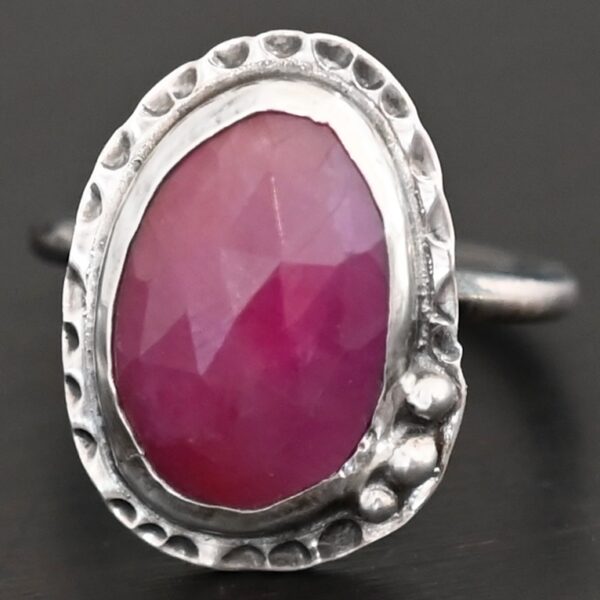 Photo of "Everything Is Rosy" Ring. Made out of sterling silver and sapphire. The sapphire is an oval shape and a dark pink, almost magenta color. It is a rose cut sapphire that is set in silver. Going around the silver setting is more hammered sterling silver in an oval shape. There are three silver balls on the top left of the oval stone setting on the hammered silver. They are right next to one another, and curve so that they go along with the curve of the sapphire. Ring has one band. Ring is sitting against a dark background.