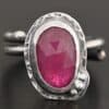 Photo of "A Whisper Of Beauty" Ring. Made out of sterling silver and sapphire. The sapphire is an oval shape and a dark pink color, with a lighter shade of pink illuminating the bottom of the ring. It is a rose cut sapphire that is set in silver. Going around the silver setting is more hammered sterling silver in an oval shape. There are three silver balls on the bottom right of the oval stone setting on the hammered silver. They are right next to one another, and curve so that they go along with the curve of the sapphire. Ring has one band that loops around to create a double band that can be adjusted to any size. Ring is sitting against a dark background.