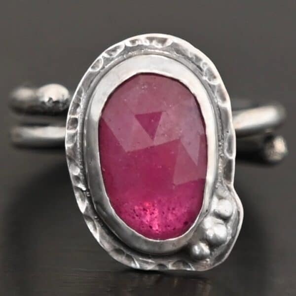 Photo of "A Whisper Of Beauty" Ring. Made out of sterling silver and sapphire. The sapphire is an oval shape and a dark pink color, with a lighter shade of pink illuminating the bottom of the ring. It is a rose cut sapphire that is set in silver. Going around the silver setting is more hammered sterling silver in an oval shape. There are three silver balls on the bottom right of the oval stone setting on the hammered silver. They are right next to one another, and curve so that they go along with the curve of the sapphire. Ring has one band that loops around to create a double band that can be adjusted to any size. Ring is sitting against a dark background.