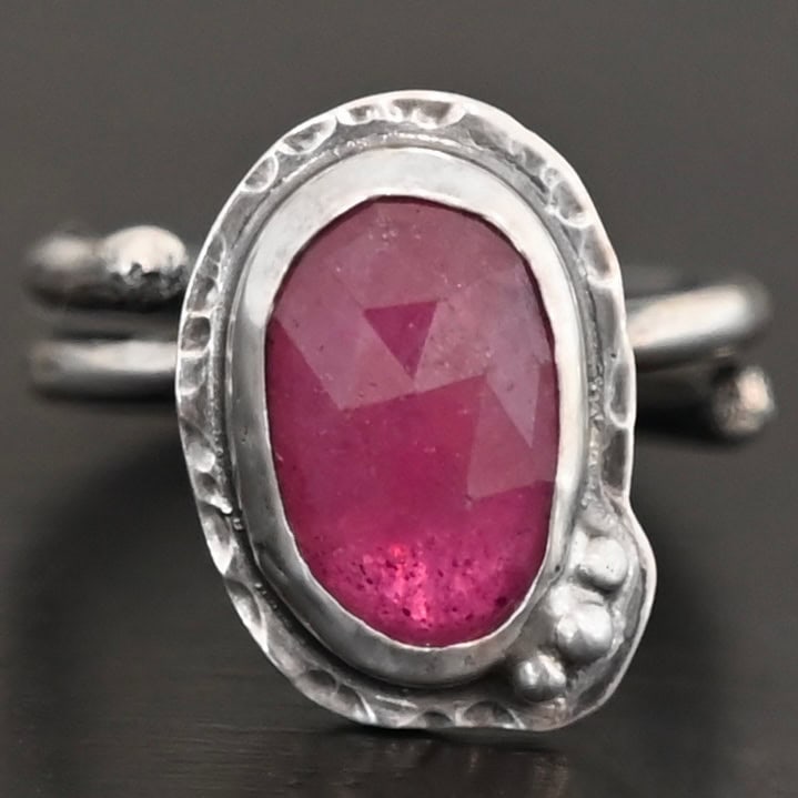 A Whisper Of Beauty Ring Photo of "A Whisper Of Beauty" Ring. Made out of sterling silver and sapphire. The sapphire is an oval shape and a dark pink color, with a lighter shade of pink illuminating the bottom of the ring. It is a rose cut sapphire that is set in silver. Going around the silver setting is more hammered sterling silver in an oval shape. There are three silver balls on the bottom right of the oval stone setting on the hammered silver. They are right next to one another, and curve so that they go along with the curve of the sapphire. Ring has one band that loops around to create a double band that can be adjusted to any size. Ring is sitting against a dark background.