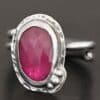 Photo of "A Whisper Of Beauty" Ring. Made out of sterling silver and sapphire. The sapphire is an oval shape and a dark pink color, with a lighter shade of pink illuminating the bottom of the ring. It is a rose cut sapphire that is set in silver. Going around the silver setting is more hammered sterling silver in an oval shape. There are three silver balls on the bottom right of the oval stone setting on the hammered silver. They are right next to one another, and curve so that they go along with the curve of the sapphire. Ring has one band that loops around to create a double band that can be adjusted to any size. Ring is sitting against a dark background.