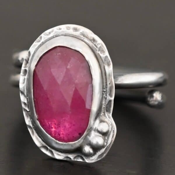 Photo of "A Whisper Of Beauty" Ring. Made out of sterling silver and sapphire. The sapphire is an oval shape and a dark pink color, with a lighter shade of pink illuminating the bottom of the ring. It is a rose cut sapphire that is set in silver. Going around the silver setting is more hammered sterling silver in an oval shape. There are three silver balls on the bottom right of the oval stone setting on the hammered silver. They are right next to one another, and curve so that they go along with the curve of the sapphire. Ring has one band that loops around to create a double band that can be adjusted to any size. Ring is sitting against a dark background.