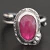 Photo of "A Whisper Of Beauty" Ring. Made out of sterling silver and sapphire. The sapphire is an oval shape and a dark pink color, with a lighter shade of pink illuminating the bottom of the ring. It is a rose cut sapphire that is set in silver. Going around the silver setting is more hammered sterling silver in an oval shape. There are three silver balls on the bottom right of the oval stone setting on the hammered silver. They are right next to one another, and curve so that they go along with the curve of the sapphire. Ring has one band that loops around to create a double band that can be adjusted to any size. Ring is sitting against a dark background.