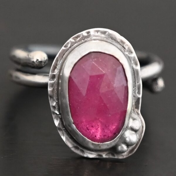 Photo of "A Whisper Of Beauty" Ring. Made out of sterling silver and sapphire. The sapphire is an oval shape and a dark pink color, with a lighter shade of pink illuminating the bottom of the ring. It is a rose cut sapphire that is set in silver. Going around the silver setting is more hammered sterling silver in an oval shape. There are three silver balls on the bottom right of the oval stone setting on the hammered silver. They are right next to one another, and curve so that they go along with the curve of the sapphire. Ring has one band that loops around to create a double band that can be adjusted to any size. Ring is sitting against a dark background.