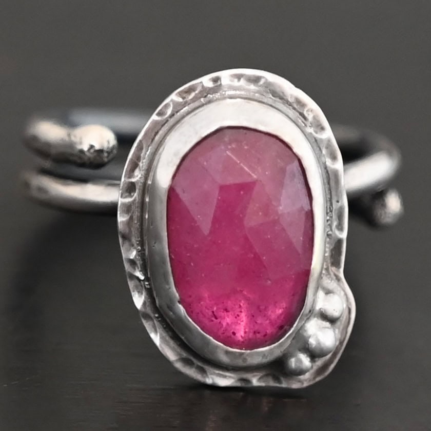 A Whisper Of Beauty Ring Photo of "A Whisper Of Beauty" Ring. Made out of sterling silver and sapphire. The sapphire is an oval shape and a dark pink color, with a lighter shade of pink illuminating the bottom of the ring. It is a rose cut sapphire that is set in silver. Going around the silver setting is more hammered sterling silver in an oval shape. There are three silver balls on the bottom right of the oval stone setting on the hammered silver. They are right next to one another, and curve so that they go along with the curve of the sapphire. Ring has one band that loops around to create a double band that can be adjusted to any size. Ring is sitting against a dark background.