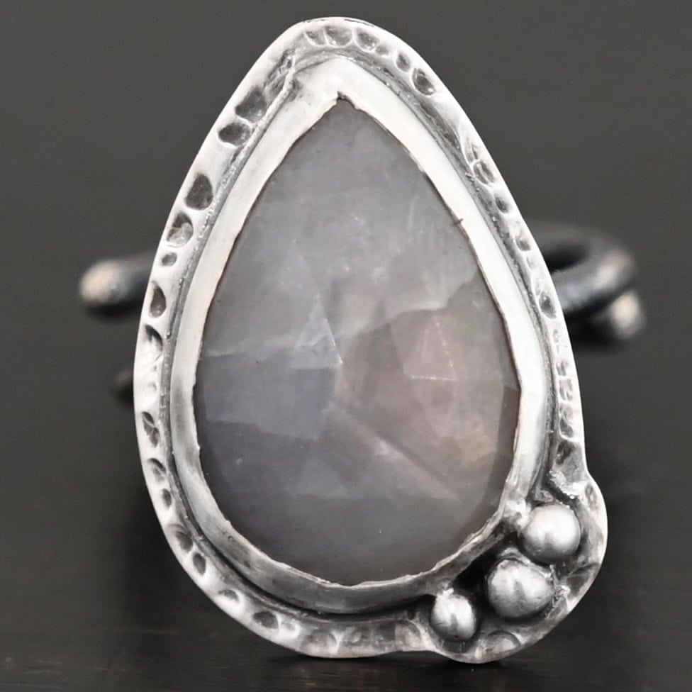 Close up of the "Peaceful Way" Ring. Made out of sterling silver and sapphire. The sapphire is a teardrop shape and rose cut. It is a dark gray color that shines because of the way it is cut. The sapphire has sterling silver going around the edge of it, with more sterling silver circling the outside of the setting that is hammered. Sitting on the hammered sterling silver at the bottom of the ring on the right side are three silver balls that are set in a slightly curved line to go along the curve of the ring. Ring is sitting against a dark background.
