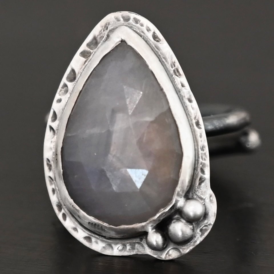 Close up of the "Peaceful Way" Ring. Made out of sterling silver and sapphire. The sapphire is a teardrop shape and rose cut. It is a dark gray color that shines because of the way it is cut. The sapphire has sterling silver going around the edge of it, with more sterling silver circling the outside of the setting that is hammered. Sitting on the hammered sterling silver at the bottom of the ring on the right side are three silver balls that are set in a slightly curved line to go along the curve of the ring. Ring is sitting against a dark background.