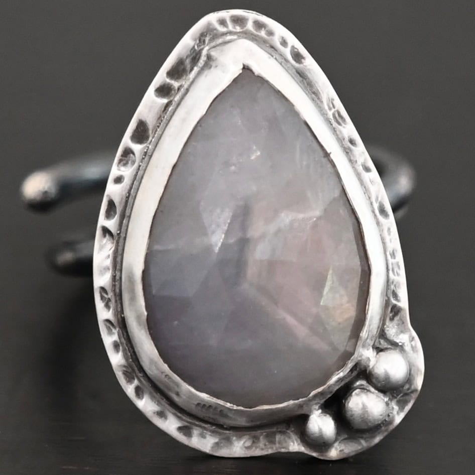 Close up of the "Peaceful Way" Ring. Made out of sterling silver and sapphire. The sapphire is a teardrop shape and rose cut. It is a dark gray color that shines because of the way it is cut. The sapphire has sterling silver going around the edge of it, with more sterling silver circling the outside of the setting that is hammered. Sitting on the hammered sterling silver at the bottom of the ring on the right side are three silver balls that are set in a slightly curved line to go along the curve of the ring. Ring is sitting against a dark background.