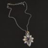 Photo of the "Arendelle" Necklace. Made out of sterling silver, gold, and tourmaline. There are three tourmalines, each a blue color. The top one is a light blue, and the middle one is a deep royal blue color. The bottom is a dark blue color as well, but not the same as the middle. Pendant is in the shape of a flower with the stones in the center going up and down. There are two layers of sterling silver, each hammered and oxidized. There are three leaf shapes on the top, one in the center and one on each side pointing opposite ways. There is a leaf on each side in the center of the necklace. There are three more leaves on the bottom of the necklace, one in the center and again, one on each side of the center one pointing opposite ways. There are gold dots around the necklace as well. There are fourteen dots and two hammered dots. Seven of the dots are on the left side. They are in groups of three. Three are on the top of the center stone and three are on the bottom. Two dots are in the middle, one that is hammered and one that is not. The hammered one is closer to the stone, and the one that is not is on the side of that one. The other seven are on the right side and follow the same pattern as the left side. There is a loop on the back of the necklace that is connected to three jump rings that are connected to the chain of the necklace. Necklace is laying against a dark background.