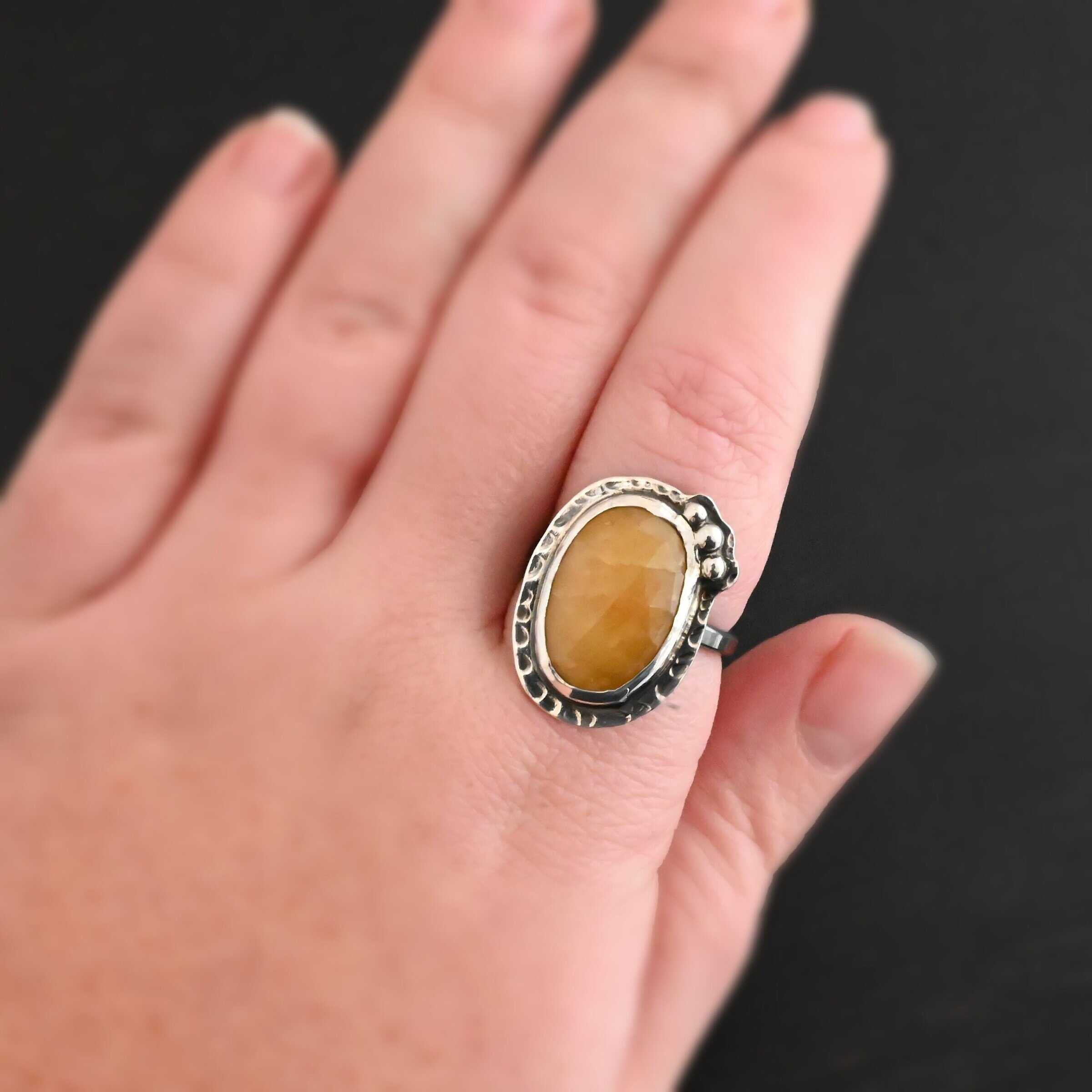 Marigold Ring Close up of the "Marigold" Ring. Made out of sterling silver and sapphire. The sapphire is an oval shape and rosecut, and it is a dark yellow color. In the bottom left corner on the hammered sterling silver surrounding the ring are three silver balls. They are slightly curved and set next to one another so that they follow the curve of the sapphire. Ring is worn on a models middle finger to demonstrate size.