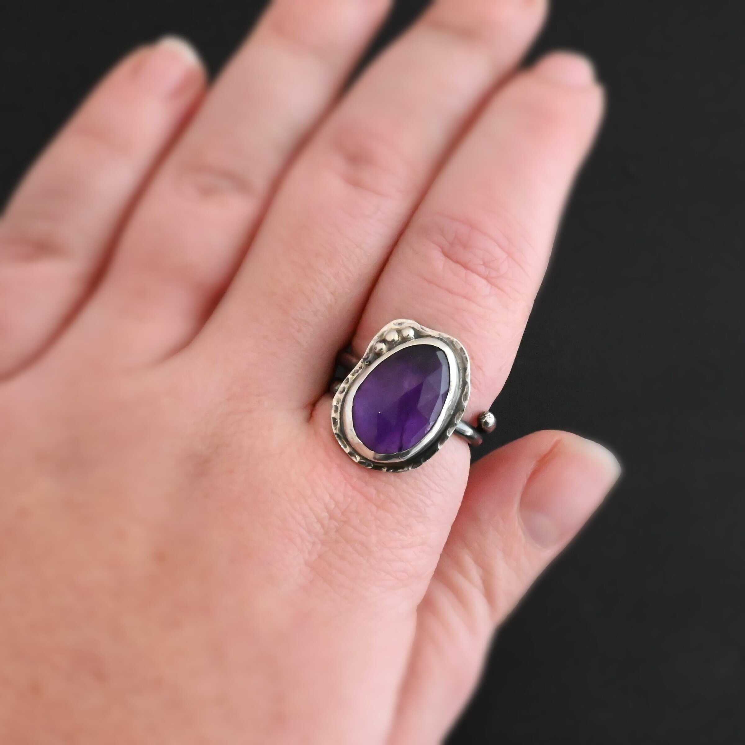 Photo of the "Let It Blossom" Ring. Ring is made out of sterling silver and amethyst. The amethyst is an oval shape. It is a dark purple color. It is set in sterling silver. There is hammered sterling silver around the stone setting as well in an oval shape. On the hammered sterling silver are three silver balls on the left side of the amethyst. They are right next to one another and follow the curve of the amethyst. Ring is worn on a models middle finger to demonstrate size.
