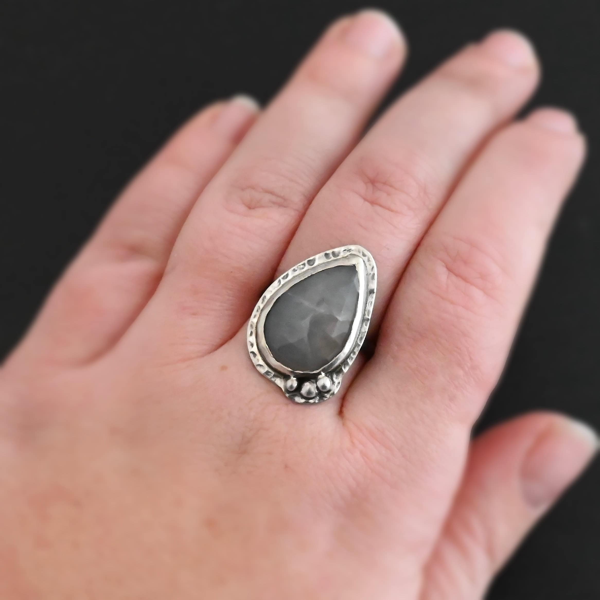 Close up of the "Peaceful Way" Ring. Made out of sterling silver and sapphire. The sapphire is a teardrop shape and rose cut. It is a dark gray color that shines because of the way it is cut. The sapphire has sterling silver going around the edge of it, with more sterling silver circling the outside of the setting that is hammered. Sitting on the hammered sterling silver at the bottom of the ring on the right side are three silver balls that are set in a slightly curved line to go along the curve of the ring. Ring is worn by a model on their ring finger to demonstrate size.