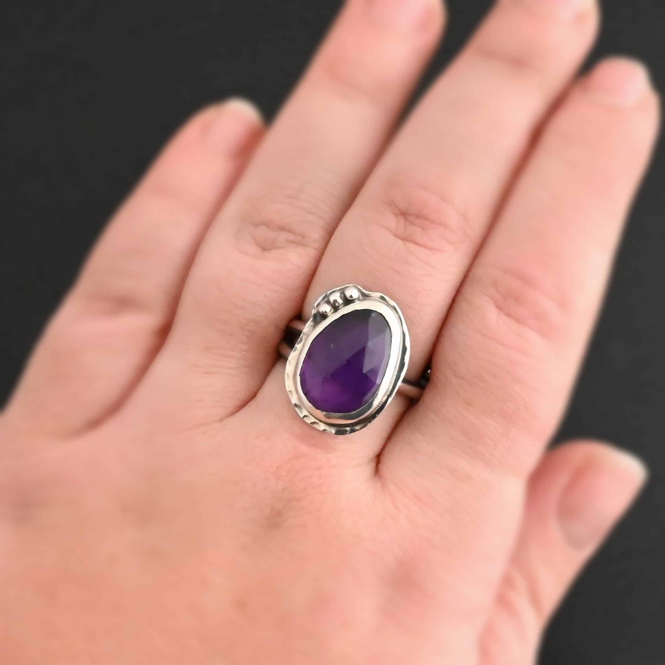 Photo of the "A Life Worthwhile" Ring. Ring is made out of sterling silver and amethyst. The amethyst is an oval shape. It is a dark purple color. It is set in sterling silver. There is hammered sterling silver around the stone setting as well in an oval shape. On the hammered sterling silver are three silver balls on the bottom right side of the amethyst. They are right next to one another and follow the curve of the amethyst. Ring has one band that does a loop around so that it looks like two bands. Ring is worn on a models middle finger to demonstrate size.