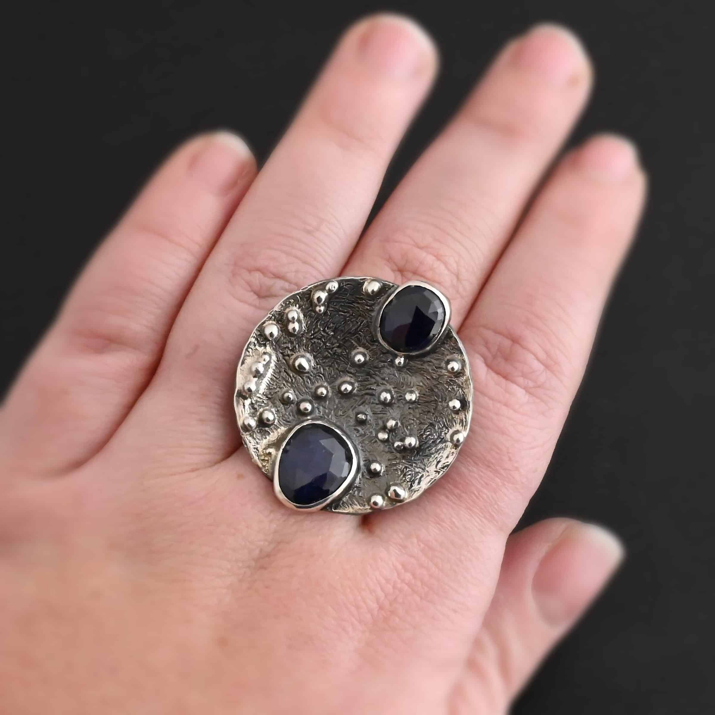 Follow The Stars Ring New Close up of the "Follow The Stars" Ring. Made out of sterling silver and sapphires. There are two sapphires, both circle shaped and rose cut. The ring is a large circle disk with silver balls scattered all over the circle ring. The sapphires are both dark blue. One of them is on the top of the ring, and sticks up a bit above the circle. The other sapphire is on the bottom of the ring, and again, sticks out below the circle. Ring is worn by a model on their pointer finger to demonstrate size of the ring.