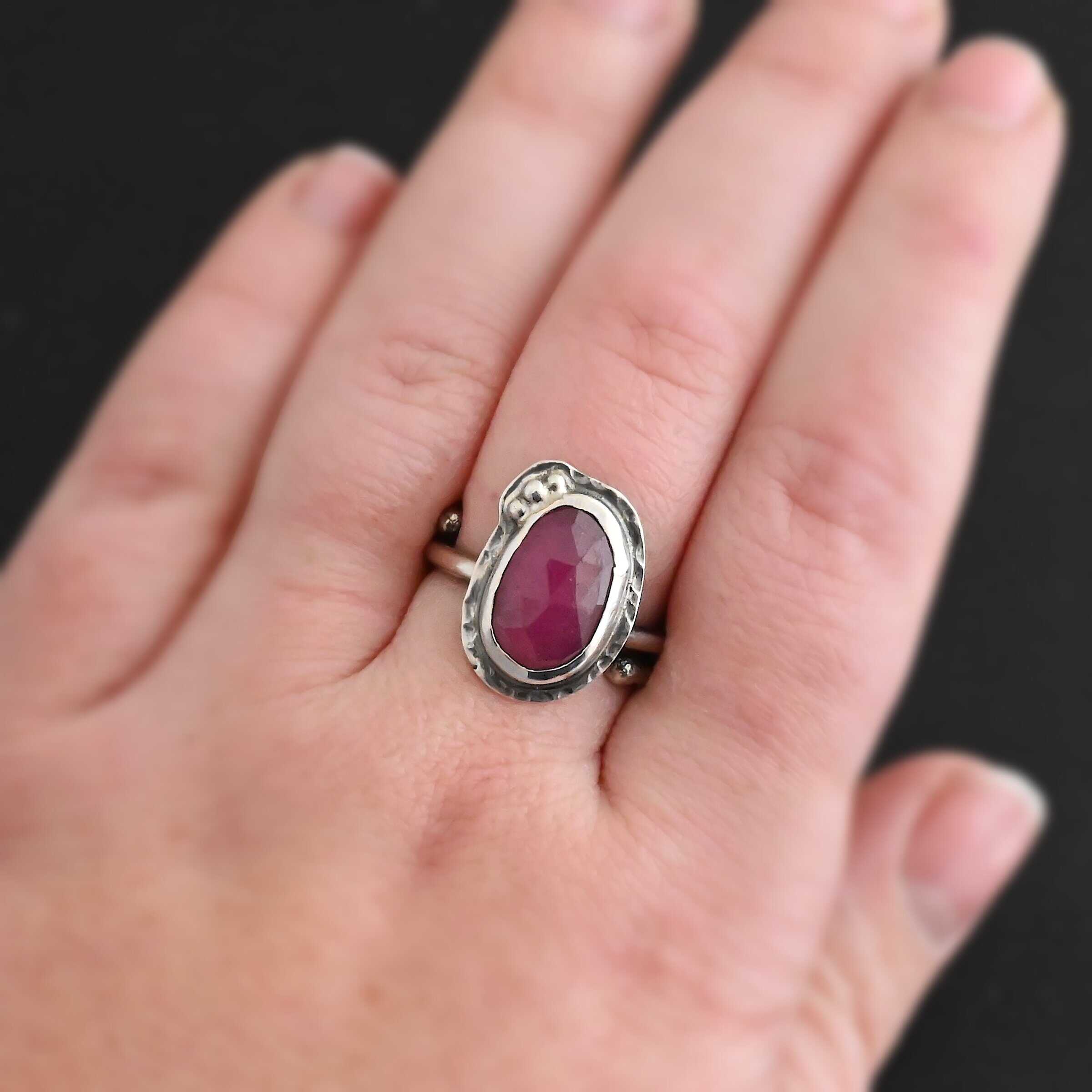 A Whisper Of Beauty Ring New Photo of "A Whisper Of Beauty" Ring. Made out of sterling silver and sapphire. The sapphire is an oval shape and a dark pink color, with a lighter shade of pink illuminating the bottom of the ring. It is a rose cut sapphire that is set in silver. Going around the silver setting is more hammered sterling silver in an oval shape. There are three silver balls on the bottom right of the oval stone setting on the hammered silver. They are right next to one another, and curve so that they go along with the curve of the sapphire. Ring has one band that loops around to create a double band that can be adjusted to any size. Ring is worn on a models middle finger to demonstrate size.