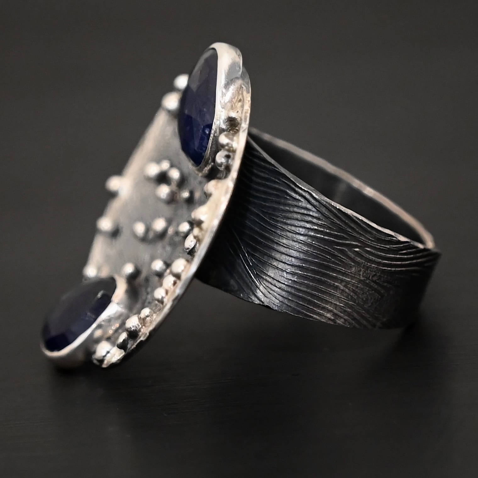 Follow The Stars Ring Close up of the "Follow The Stars" Ring. Made out of sterling silver and sapphires. There are two sapphires, both circle shaped and rose cut. The ring is a large circle disk with silver balls scattered all over the circle ring. The sapphires are both dark blue. One of them is on the top of the ring, and sticks up a bit above the circle. The other sapphire is on the bottom of the ring, and again, sticks out below the circle. The ring band is engraved with wavy lines all around it. Ring is sitting against a dark background.