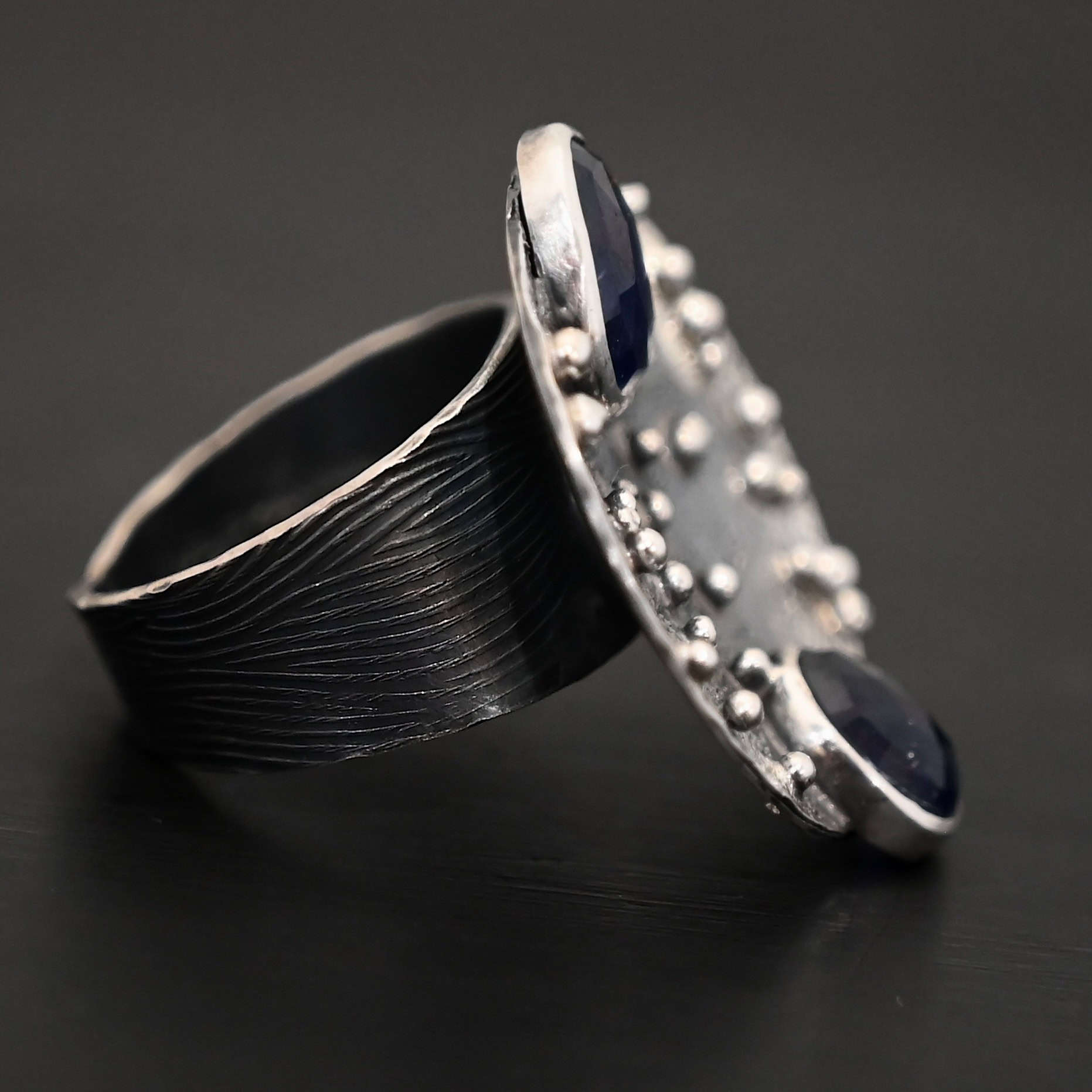 Follow The Stars Ring Close up of the "Follow The Stars" Ring. Made out of sterling silver and sapphires. There are two sapphires, both circle shaped and rose cut. The ring is a large circle disk with silver balls scattered all over the circle ring. The sapphires are both dark blue. One of them is on the top of the ring, and sticks up a bit above the circle. The other sapphire is on the bottom of the ring, and again, sticks out below the circle. The ring band is engraved with wavy lines all around it. Ring is sitting against a dark background.
