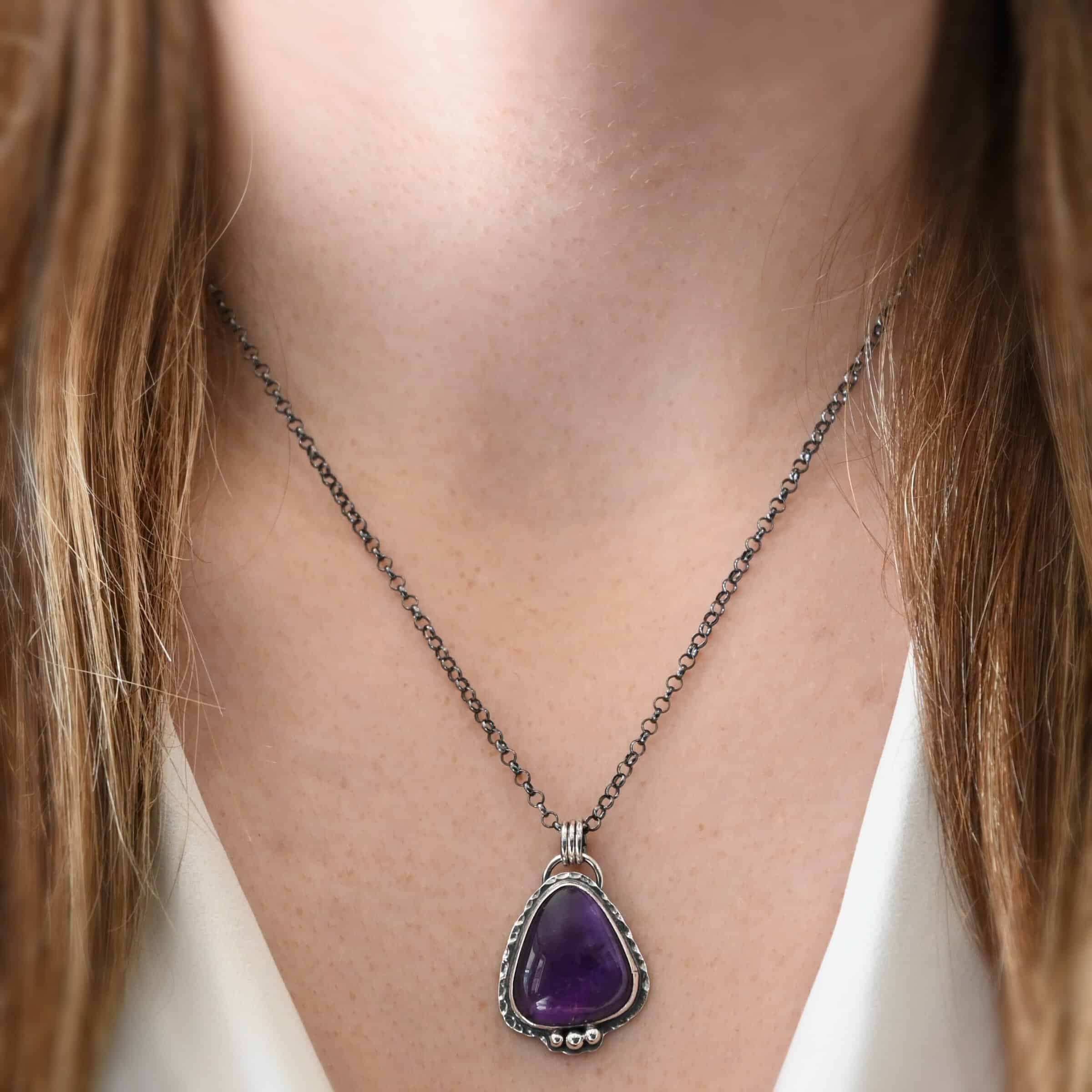 Ought To Do Good Necklace Photo of the "Ought To Do Good" Necklace. Made out of sterling silver and amethyst. The amethyst is a triangle shape but with curved points. The bottom of the amethyst has three silver dots in a straight line that go across the flat bottom. The rest of the silver setting is hammered. There is a loop on the back with three jump rings that are attached to the chain of the necklace. Necklace is worn by a model at 18 inches long to demonstrate size of the pendant.