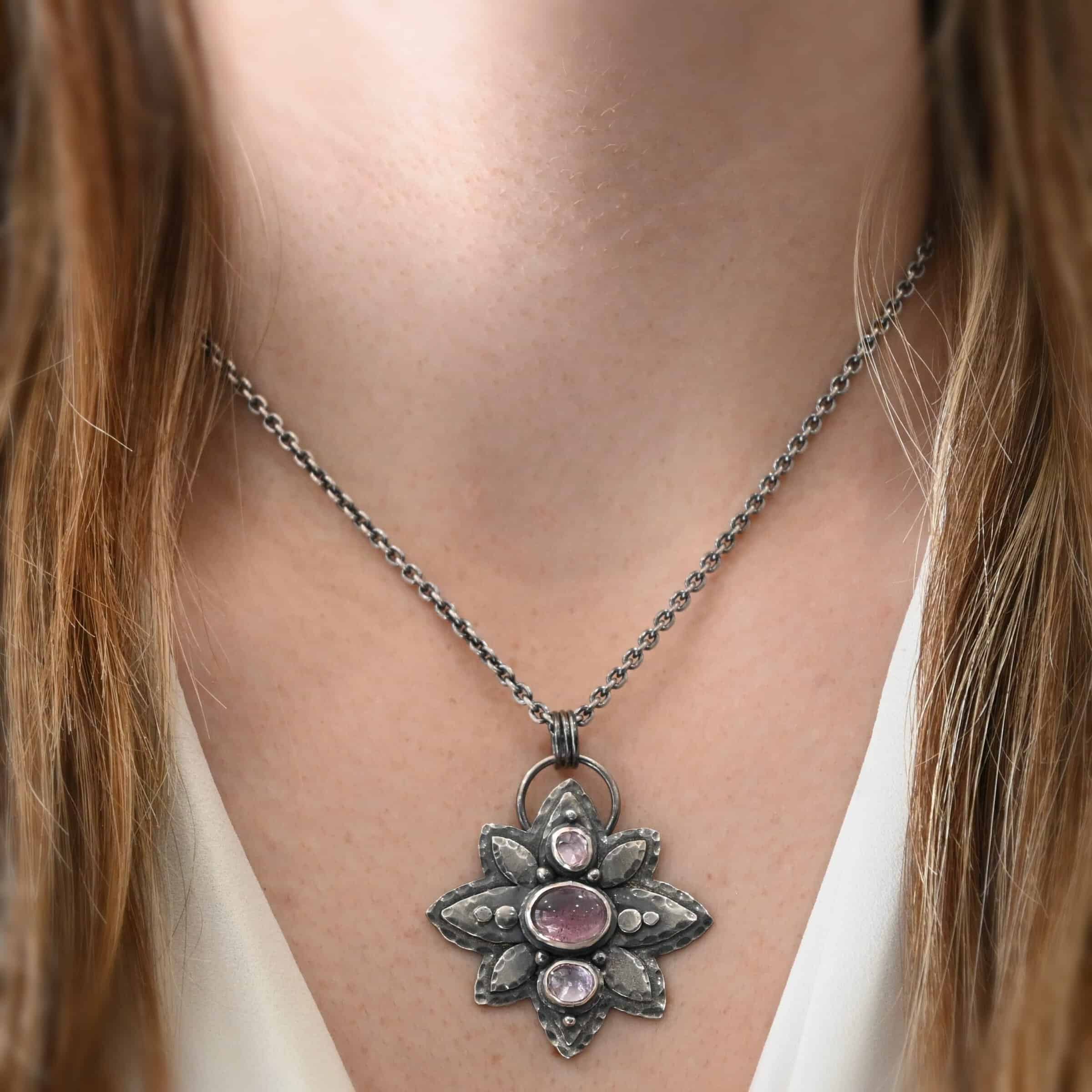 Iduna Necklace Photo of the "Iduna" Necklace. Made out of sterling silver and tourmaline. There are three tourmalines, each a purple color. The top and bottom are light purple, and the middle one is a dark purple. Pendant is in the shape of a flower with the stones in the center going up and down. There are two layers of sterling silver, each hammered and oxidized. There are three leaf shapes on the top, one in the center and one on each side pointing opposite ways. There is a leaf on each side in the center of the necklace. There are three more leaves on the bottom of the necklace, one in the center and again, one on each side of the center one pointing opposite ways. There are silver dots around the necklace as well. One is on the top stone, and two beneath it on each side of the stone. There are two more silver dots on the bottom of the middle stone on each side of it. There is one more silver dot on the bottom stone. There are four silver dots in the center of the necklace, two on each of the center leaves. These dots are hammered so that they are flat. There is a loop on the back of the necklace that is attached to jump rings that are hung on the chain of the necklace. Necklace is worn at 17 inches long by a model to demonstrate size of the pendant.