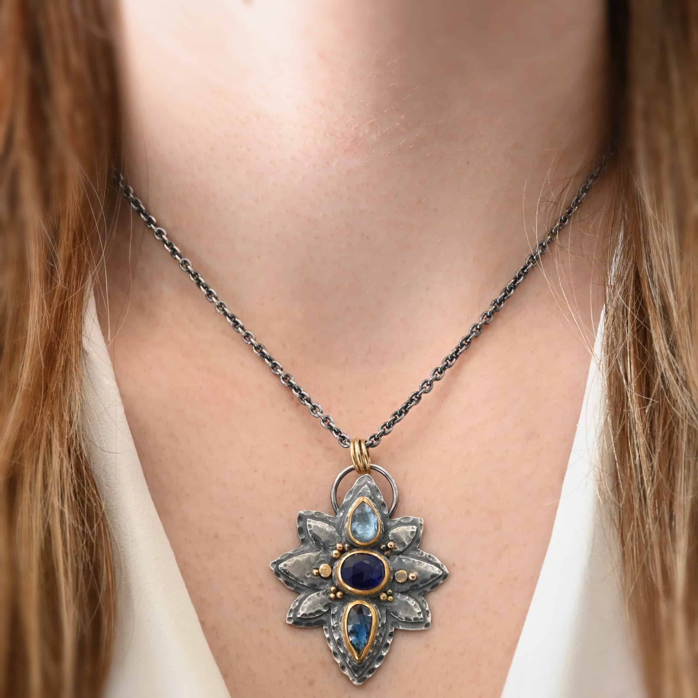 Arendelle Necklace Photo of the "Arendelle" Necklace. Made out of sterling silver, gold, and tourmaline. There are three tourmalines, each a blue color. The top one is a light blue, and the middle one is a deep royal blue color. The bottom is a dark blue color as well, but not the same as the middle. Pendant is in the shape of a flower with the stones in the center going up and down. There are two layers of sterling silver, each hammered and oxidized. There are three leaf shapes on the top, one in the center and one on each side pointing opposite ways. There is a leaf on each side in the center of the necklace. There are three more leaves on the bottom of the necklace, one in the center and again, one on each side of the center one pointing opposite ways. There are gold dots around the necklace as well. There are fourteen dots and two hammered dots. Seven of the dots are on the left side. They are in groups of three. Three are on the top of the center stone and three are on the bottom. Two dots are in the middle, one that is hammered and one that is not. The hammered one is closer to the stone, and the one that is not is on the side of that one. The other seven are on the right side and follow the same pattern as the left side. There is a loop on the back of the necklace that is connected to three jump rings that are connected to the chain of the necklace. Necklace is worn by a model at 16 inches long to demonstrate size of the pendant.