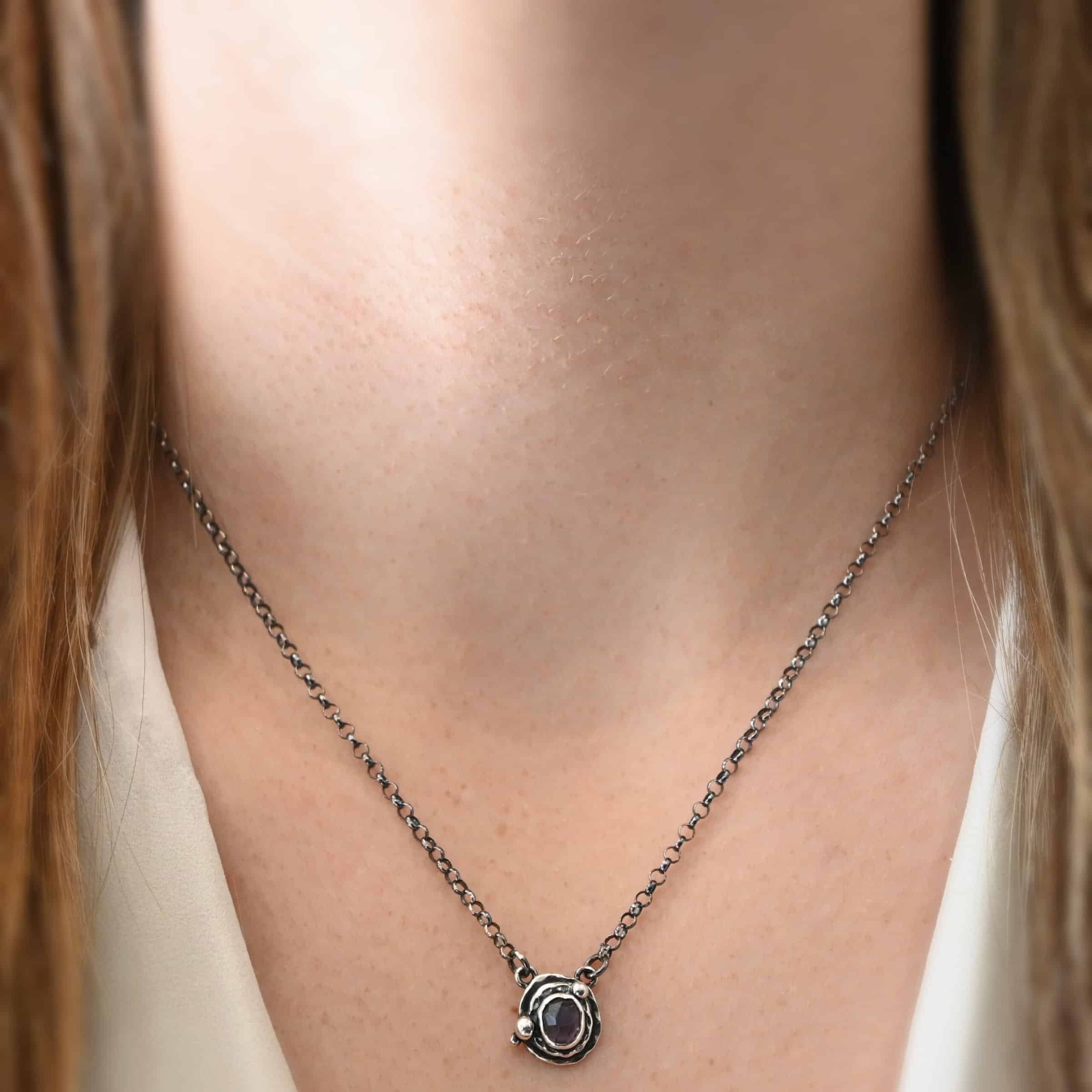 Photo of the "Vera" Necklace. Made out of sterling silver and tourmaline. The necklace has a small tourmaline in the center, of which is a deep purple color. The necklace has two layers of sterling silver, and both are hammered. The necklace also has three silver balls scattered around the pendant part of the necklace. There are two loops on the back of the pendant to connect the chain for the necklace to. The necklace is worn by a model at 18" long to demonstrate size of the pendant.