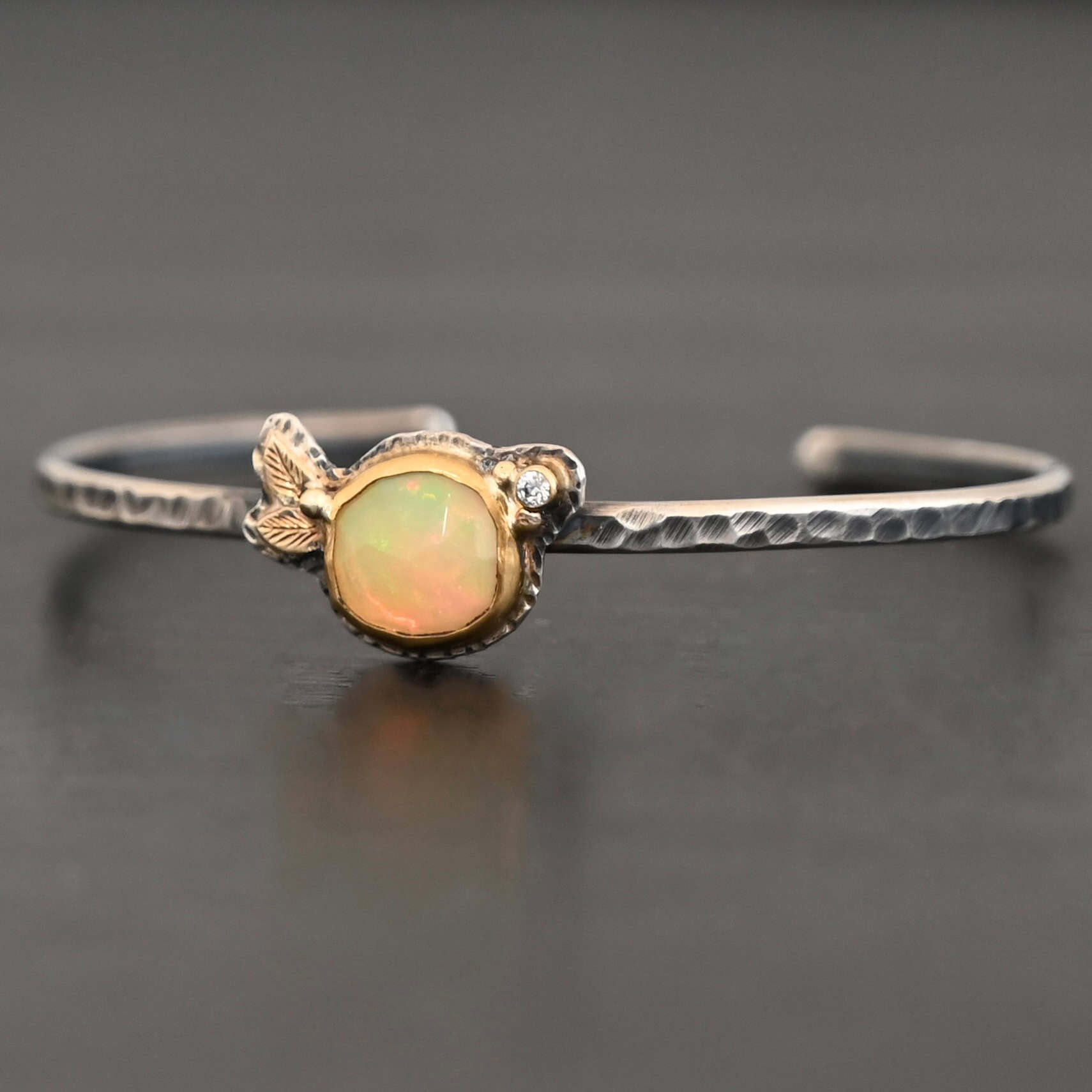 Eternity's Sunrise Cuff Photo of "Eternity's Sunrise" Cuff. Made out of sterling silver, gold, opal, and diamond. There is a circular opal in the center of the cuff. It has vibrant, but translucent red colors shining through it. There is also a light translucent green shining through it. The opal is set in gold. There are two gold leaves on the left side of the cuff, right next to each other. There is a gold dot in the center of the leaves, attached to the gold setting around the opal. There is a diamond set in gold on the right side of the opal. There are two gold dots, one on each side of the diamond. The cuff of the bracelet is made out of sterling silver, and is hammered. Bracelet is sitting against a dark background.