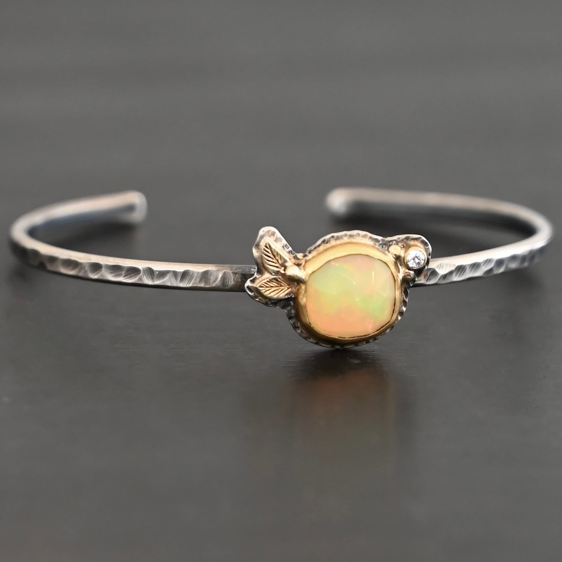 Eternity's Sunrise Cuff Photo of "Eternity's Sunrise" Cuff. Made out of sterling silver, gold, opal, and diamond. There is a circular opal in the center of the cuff. It has vibrant, but translucent red colors shining through it. There is also a light translucent green shining through it. The opal is set in gold. There are two gold leaves on the left side of the cuff, right next to each other. There is a gold dot in the center of the leaves, attached to the gold setting around the opal. There is a diamond set in gold on the right side of the opal. There are two gold dots, one on each side of the diamond. The cuff of the bracelet is made out of sterling silver, and is hammered. Bracelet is sitting against a dark background.