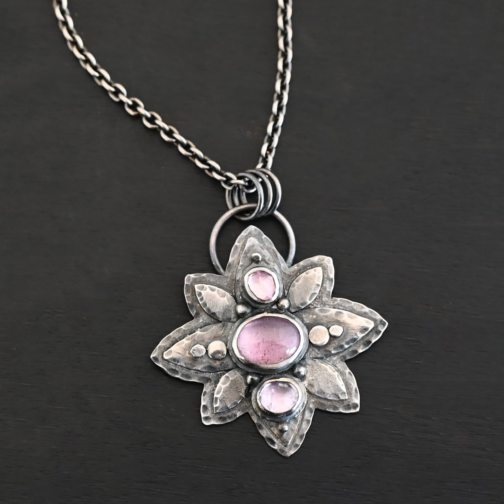 Iduna Necklace New Photo of the "Iduna" Necklace. Made out of sterling silver and tourmaline. There are three tourmalines, each a purple color. The top and bottom are light purple, and the middle one is a dark purple. Pendant is in the shape of a flower with the stones in the center going up and down. There are two layers of sterling silver, each hammered and oxidized. There are three leaf shapes on the top, one in the center and one on each side pointing opposite ways. There is a leaf on each side in the center of the necklace. There are three more leaves on the bottom of the necklace, one in the center and again, one on each side of the center one pointing opposite ways. There are silver dots around the necklace as well. One is on the top stone, and two beneath it on each side of the stone. There are two more silver dots on the bottom of the middle stone on each side of it. There is one more silver dot on the bottom stone. There are four silver dots in the center of the necklace, two on each of the center leaves. These dots are hammered so that they are flat. There is a loop on the back of the necklace that is attached to jump rings that are hung on the chain of the necklace. Necklace is laying against a dark background.