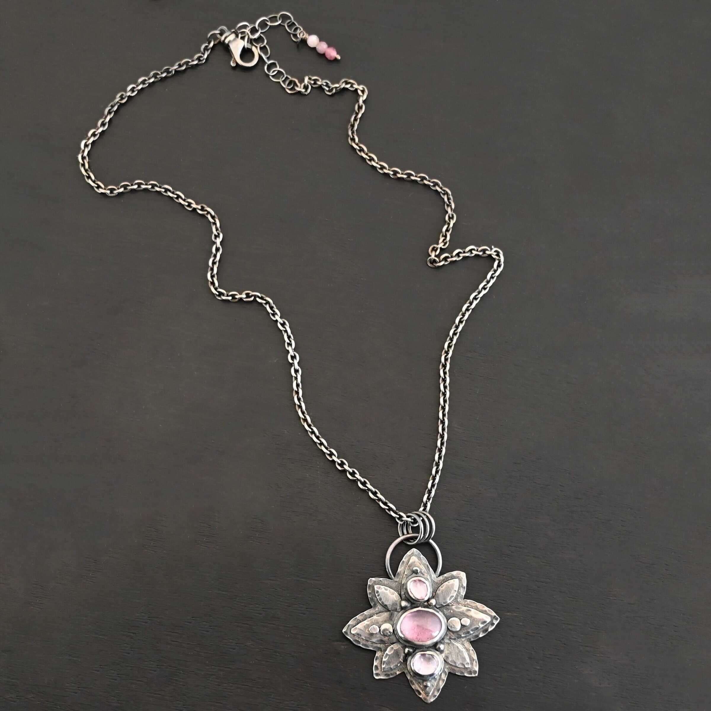 Iduna Necklace New Photo of the "Iduna" Necklace. Made out of sterling silver and tourmaline. There are three tourmalines, each a purple color. The top and bottom are light purple, and the middle one is a dark purple. Pendant is in the shape of a flower with the stones in the center going up and down. There are two layers of sterling silver, each hammered and oxidized. There are three leaf shapes on the top, one in the center and one on each side pointing opposite ways. There is a leaf on each side in the center of the necklace. There are three more leaves on the bottom of the necklace, one in the center and again, one on each side of the center one pointing opposite ways. There are silver dots around the necklace as well. One is on the top stone, and two beneath it on each side of the stone. There are two more silver dots on the bottom of the middle stone on each side of it. There is one more silver dot on the bottom stone. There are four silver dots in the center of the necklace, two on each of the center leaves. These dots are hammered so that they are flat. There is a loop on the back of the necklace that is attached to jump rings that are hung on the chain of the necklace. Necklace is laying against a dark background.