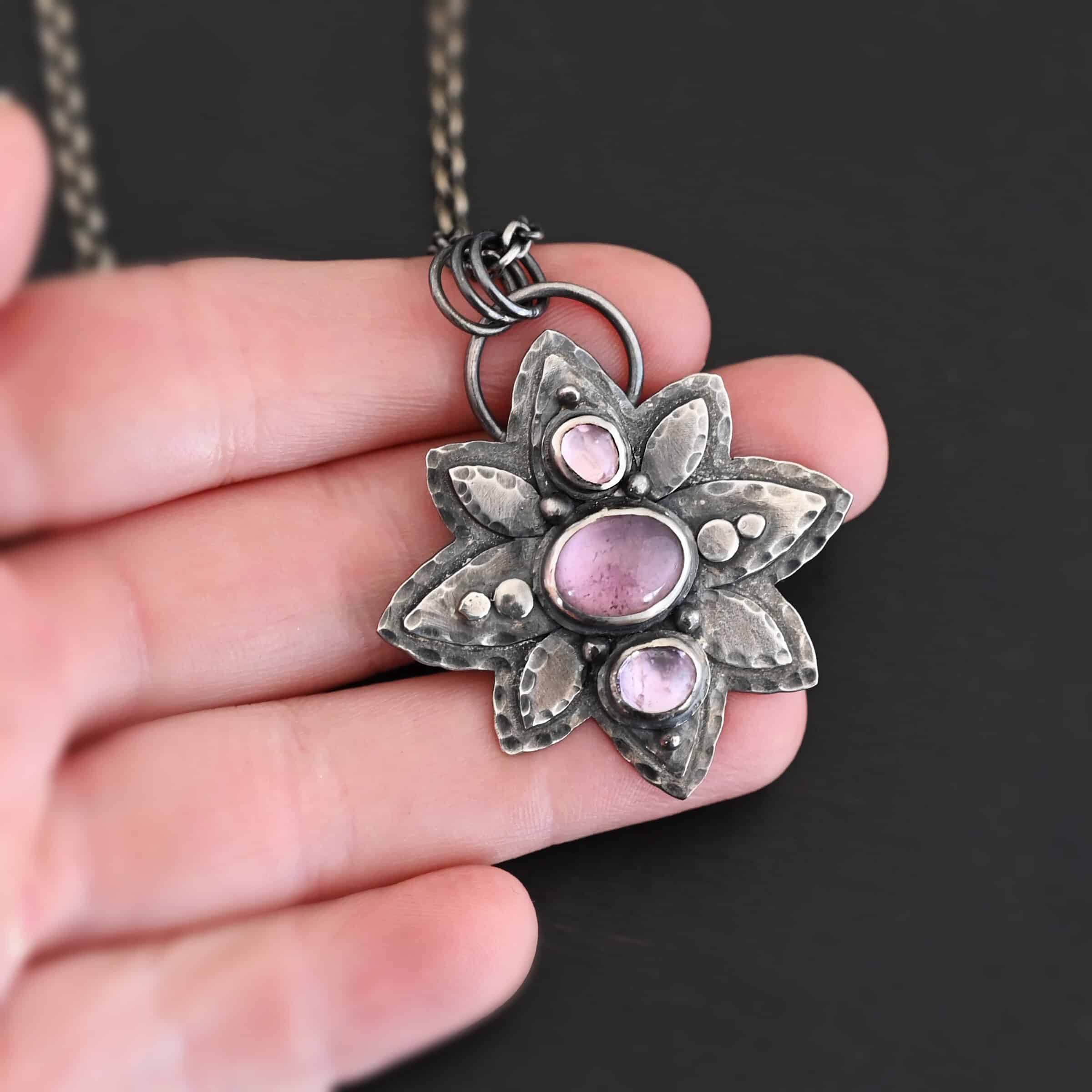 Iduna Necklace New Photo of the "Iduna" Necklace. Made out of sterling silver and tourmaline. There are three tourmalines, each a purple color. The top and bottom are light purple, and the middle one is a dark purple. Pendant is in the shape of a flower with the stones in the center going up and down. There are two layers of sterling silver, each hammered and oxidized. There are three leaf shapes on the top, one in the center and one on each side pointing opposite ways. There is a leaf on each side in the center of the necklace. There are three more leaves on the bottom of the necklace, one in the center and again, one on each side of the center one pointing opposite ways. There are silver dots around the necklace as well. One is on the top stone, and two beneath it on each side of the stone. There are two more silver dots on the bottom of the middle stone on each side of it. There is one more silver dot on the bottom stone. There are four silver dots in the center of the necklace, two on each of the center leaves. These dots are hammered so that they are flat. There is a loop on the back of the necklace that is attached to jump rings that are hung on the chain of the necklace. Necklace is laying against the fingertips of a hand.