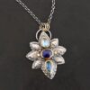 Photo of the "Arendelle" Necklace. Made out of sterling silver, gold, and tourmaline. There are three tourmalines, each a blue color. The top one is a light blue, and the middle one is a deep royal blue color. The bottom is a dark blue color as well, but not the same as the middle. Pendant is in the shape of a flower with the stones in the center going up and down. There are two layers of sterling silver, each hammered and oxidized. There are three leaf shapes on the top, one in the center and one on each side pointing opposite ways. There is a leaf on each side in the center of the necklace. There are three more leaves on the bottom of the necklace, one in the center and again, one on each side of the center one pointing opposite ways. There are gold dots around the necklace as well. There are fourteen dots and two hammered dots. Seven of the dots are on the left side. They are in groups of three. Three are on the top of the center stone and three are on the bottom. Two dots are in the middle, one that is hammered and one that is not. The hammered one is closer to the stone, and the one that is not is on the side of that one. The other seven are on the right side and follow the same pattern as the left side. There is a loop on the back of the necklace that is connected to three jump rings that are connected to the chain of the necklace. Necklace is laying against a dark background.