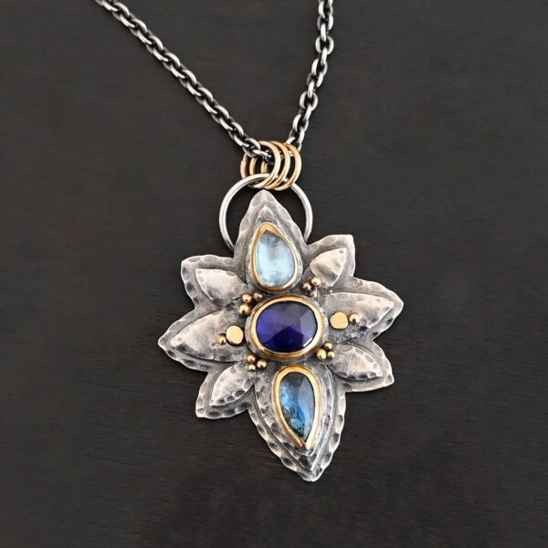 Photo of the "Arendelle" Necklace. Made out of sterling silver, gold, and tourmaline. There are three tourmalines, each a blue color. The top one is a light blue, and the middle one is a deep royal blue color. The bottom is a dark blue color as well, but not the same as the middle. Pendant is in the shape of a flower with the stones in the center going up and down. There are two layers of sterling silver, each hammered and oxidized. There are three leaf shapes on the top, one in the center and one on each side pointing opposite ways. There is a leaf on each side in the center of the necklace. There are three more leaves on the bottom of the necklace, one in the center and again, one on each side of the center one pointing opposite ways. There are gold dots around the necklace as well. There are fourteen dots and two hammered dots. Seven of the dots are on the left side. They are in groups of three. Three are on the top of the center stone and three are on the bottom. Two dots are in the middle, one that is hammered and one that is not. The hammered one is closer to the stone, and the one that is not is on the side of that one. The other seven are on the right side and follow the same pattern as the left side. There is a loop on the back of the necklace that is connected to three jump rings that are connected to the chain of the necklace. Necklace is laying against a dark background.